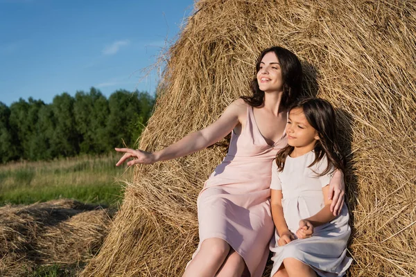 Brunette woman looking away and pointing with hand on haystack near daughter — Stockfoto