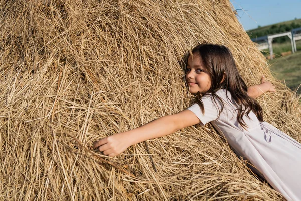 Pleased brunette girl embracing haystack and looking at camera — Stock Photo