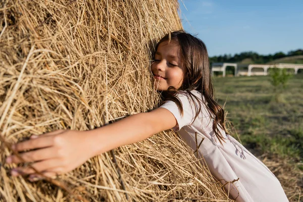 Smiling girl with closed eyes embracing haystack in meadow — Photo de stock