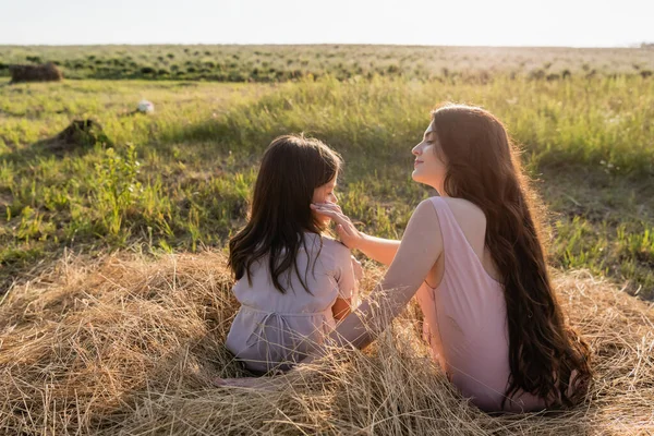 Brunette woman touching face of daughter while sitting on haystack in field — Stock Photo