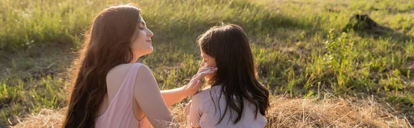 Woman with long hair touching face of daughter while sitting in meadow, banner - foto de stock