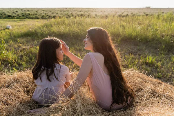 Woman with long hair sitting near daughter in field and touching her face — Stock Photo