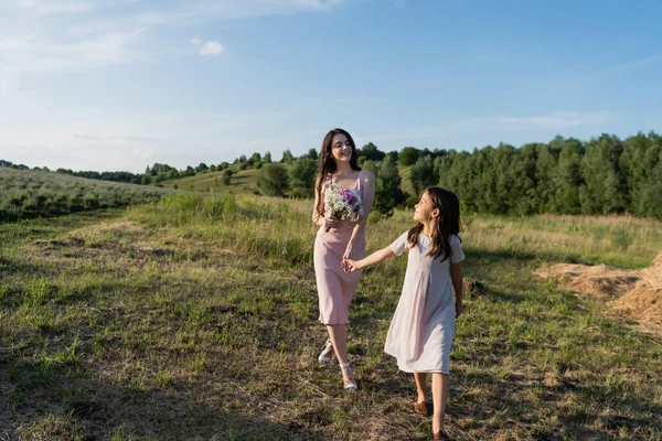 Woman with bouquet holding hands with daughter while walking in field - foto de stock