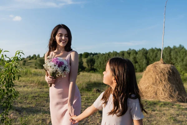 Cheerful woman with flowers holding hands with daughter in meadow on summer day — Stock Photo