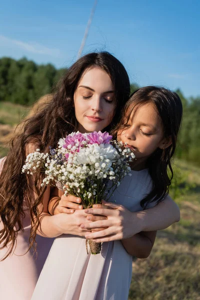 Woman and child with closed eyes smelling aromatic flowers in blurred meadow - foto de stock