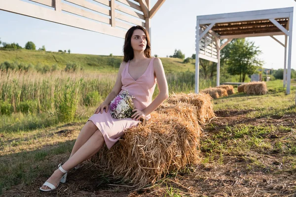 Full length of woman in summer dress sitting on haystack and looking away — Stock Photo