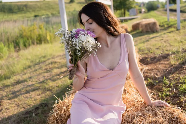Brunette woman with closed eyes sitting on haystack and smelling bouquet — Stockfoto