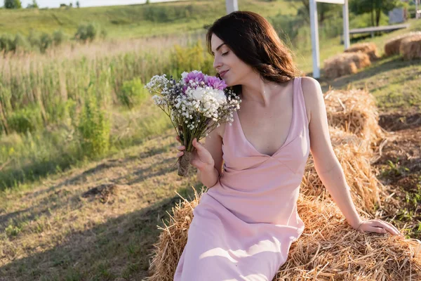 Pretty woman smelling aromatic bouquet while sitting on haystack — Fotografia de Stock