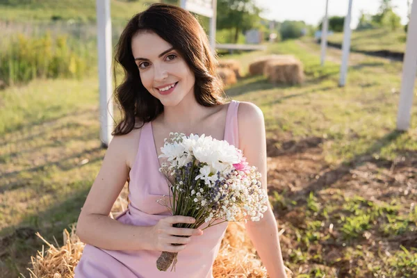Cheerful brunette woman with bouquet smiling at camera in countryside — Fotografia de Stock