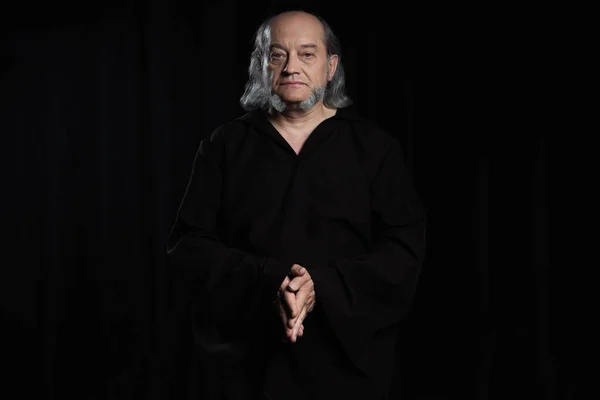 Medieval monk in dark cassock standing with folded hands and looking at camera isolated on black — Stock Photo