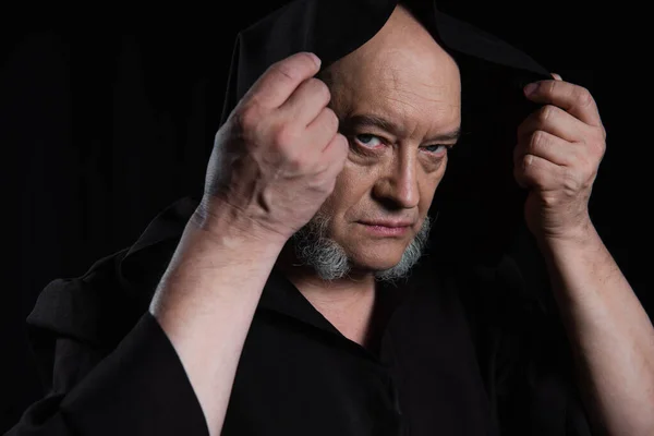 Mysterious monk in hooded cassock looking at camera isolated on black — Stock Photo