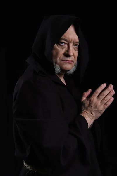 Strict monk in hooded cassock looking at camera while standing with praying hands isolated on black — Stock Photo