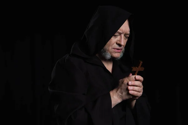 Senior monk in hood looking at wooden cross while praying isolated on black — Stock Photo