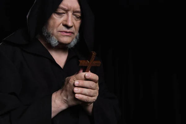 Senior priest in dark hood looking at holy crucifix isolated on black — Stock Photo