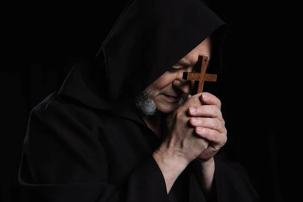Senior monk in hooded cassock praying with crucifix near face isolated on black — Stock Photo