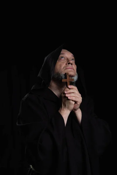 Misterioso sacerdote medieval con cruz de madera mirando hacia arriba aislado en negro - foto de stock