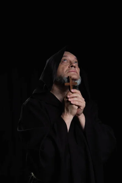 Mysterious medieval priest with wooden cross looking up isolated on black — Stock Photo