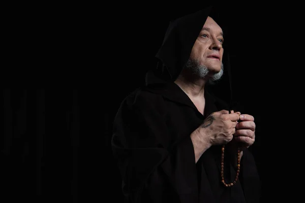 Tattooed monk in hooded cassock praying with rosary isolated on black — Stock Photo