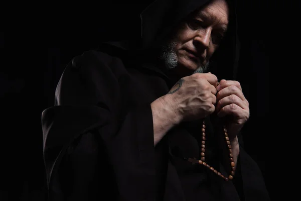 Low angle view of monk in hooded robe praying with rosary isolated on black — Stock Photo