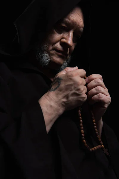 Senior tattooed monk in hood holding rosary while praying isolated on black — Stock Photo