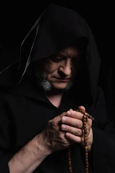 Medieval priest in hooded robe praying with rosary beads isolated on black — Stock Photo