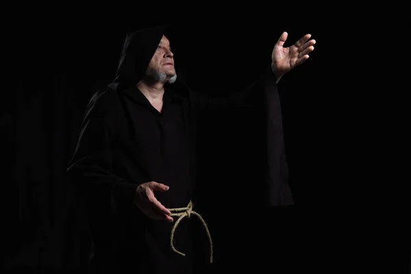 Monk in cassock with hood pointing with hand while praying isolated on black — Stock Photo