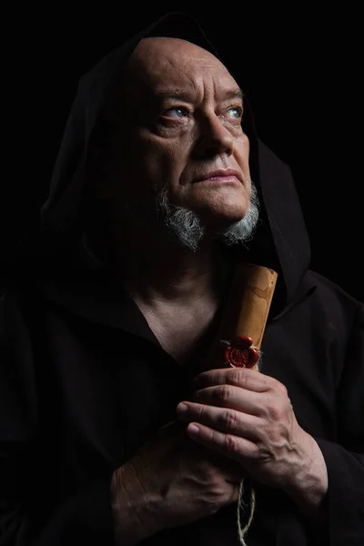 Thoughtful monk holding ancient parchment with wax seal and looking away isolated on black — Stock Photo