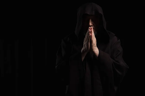 Front view of mysterious monk in hooded cassock praying isolated on black — Stock Photo