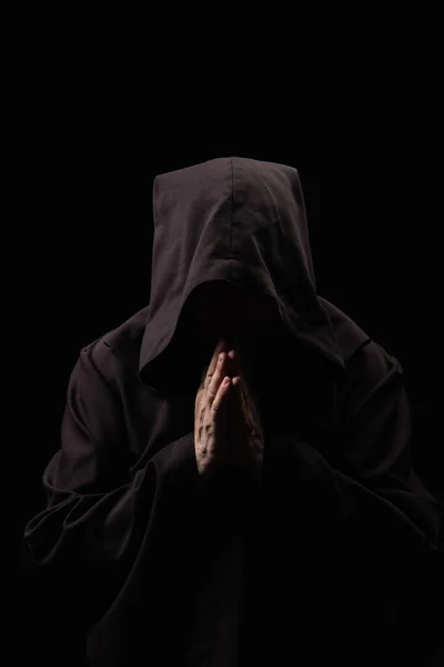 Monk in hooded robe with praying hands and obscured face isolated on black — Stock Photo