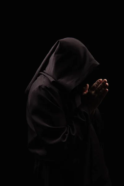 Vista lateral de monge misterioso no capuz orando isolado no preto — Fotografia de Stock