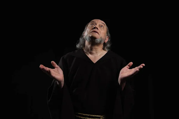 Medieval monk in dark cassock looking up while praying with outstretched hands isolated on black — Stock Photo