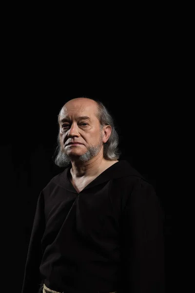 Sacerdote serio con el pelo gris y la barba mirando a la cámara aislada en negro - foto de stock