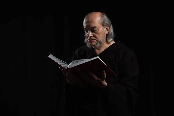 Medieval priest in dark robe reading holy bible isolated on black — Stock Photo