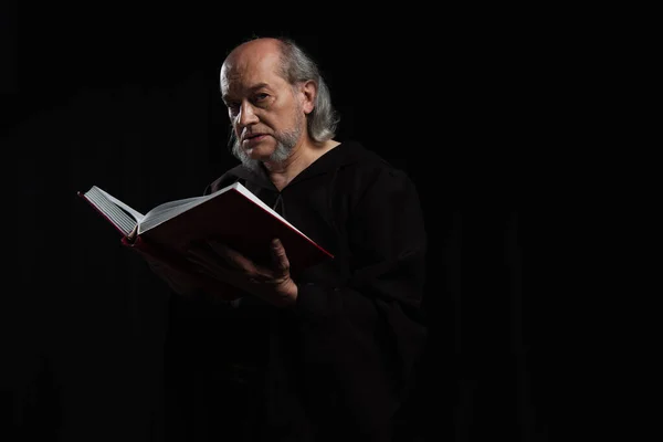 Mysterious monk in robe holding holy bible and looking at camera isolated on black — Stock Photo