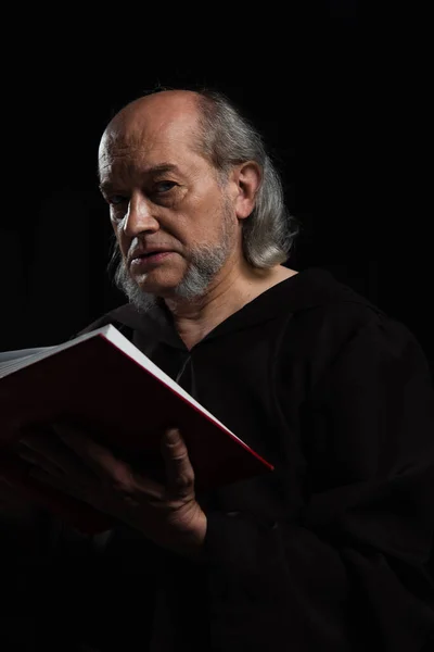 Sacerdote anziano con Sacra Bibbia guardando la macchina fotografica isolata su nero — Foto stock