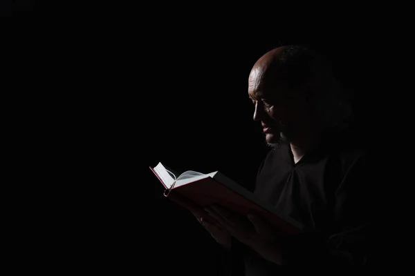 Mysterious monk in robe reading bible in darkness isolated on black — Stock Photo