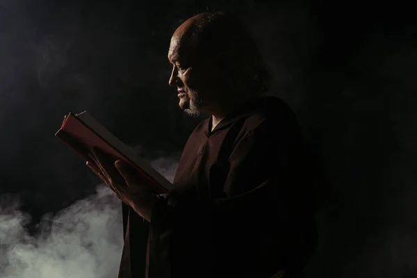 Side view of senior priest in cassock reading holy bible on black with smoke — Stock Photo