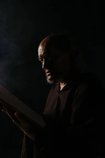 Senior medieval monk reading holy bible in darkness isolated on black — Stock Photo