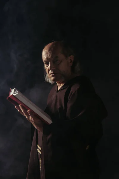 Strict senior monk looking at camera while holding bible on black with smoke — Stock Photo