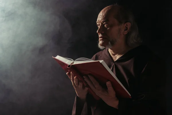 Mysterious monk with holy bible looking away on black with smoke — Stock Photo