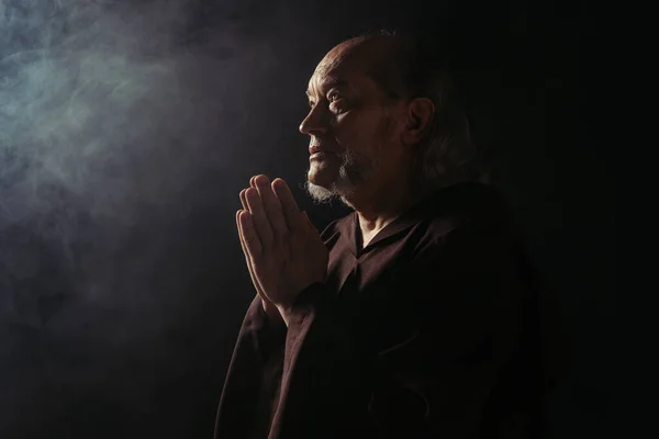 Mysterious monk in black cassock praying on dark background with smoke — Stock Photo