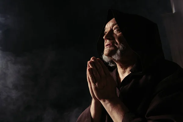 Low angle view of monk in black hooded robe praying on dark background — Stock Photo