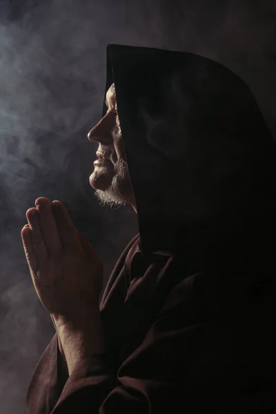 Side view of medieval monk in hooded robe praying on black with smoke — Stock Photo