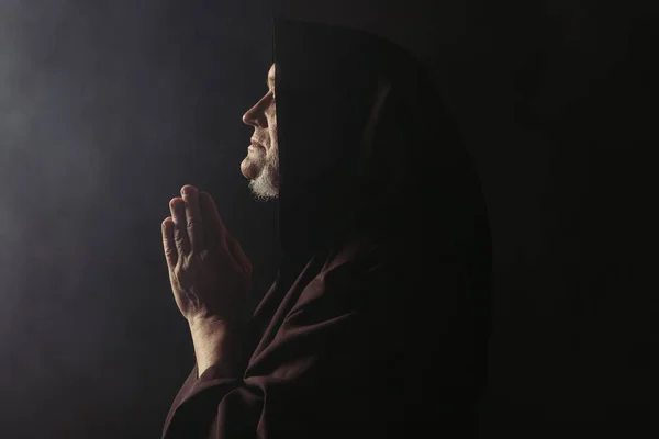 Side view of medieval monk in hooded robe praying on black with smoke — Stock Photo