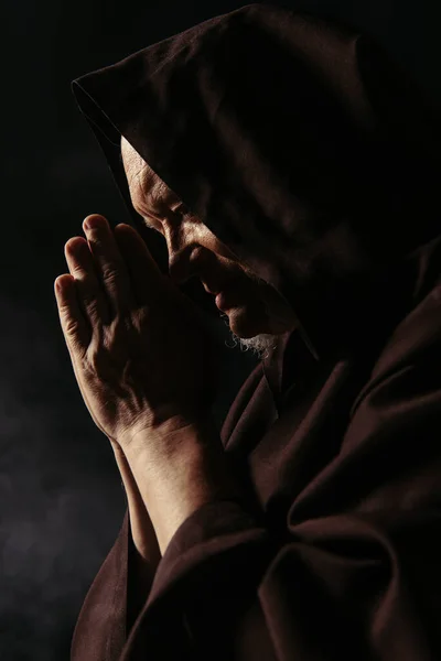 Vista lateral del sacerdote mayor con las manos en oración y en capucha negra sobre fondo oscuro - foto de stock