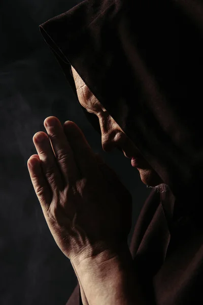 Вид збоку старшого монаха в темному капюшоні і з молитовними руками ізольовані на чорному — стокове фото