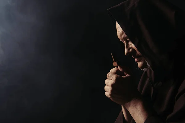 Side view of monk in hood praying with holy crucifix on black background — Stock Photo