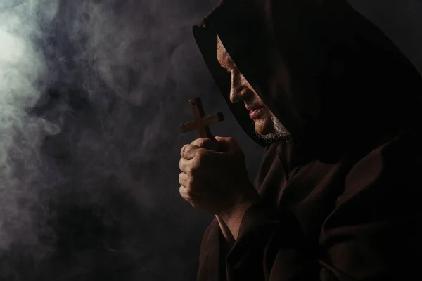 Side view of monk in robe holding holy cross while praying on black with smoke — Stock Photo