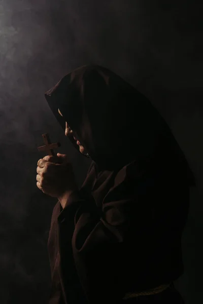 Mysterious medieval monk in hooded robe praying with crucifix on black background — Stock Photo