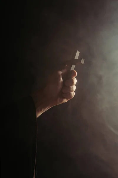 Partial view of monk with holy cross in hand on black background with smoke — Stock Photo